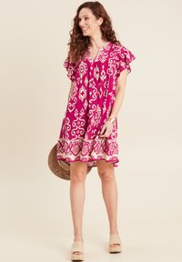 Kurzes rosafarbenes Kleid mit weißen geometrischen Mustern, fließenden Ärmeln, einem V-Ausschnitt und einem ausgestellten Saum, kombiniert mit beigen Espadrilles.