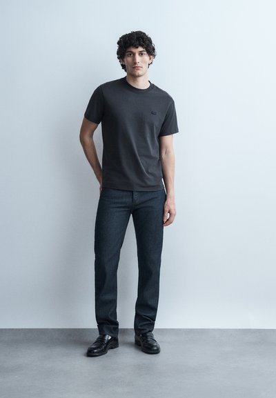 Νεαρός άνδρας με σγουρά μαλλιά που φοράει μαύρο μπλουζάκι, σκούρο τζιν και μαύρα παπούτσια, στέκεται μπροστά σε έναν απλό ανοιχτό γκρι τοίχο.