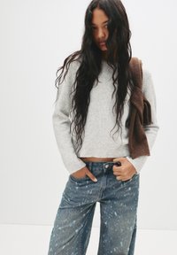 Pulover gri scurt cu o textură moale, asortat cu blugi largi de culoare albastru deschis, cu un model stropit. Accesoriu din tricot de culoare maronie purtat pe umăr.