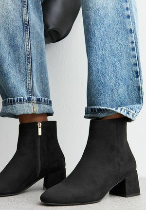 Bottes noires en daim à chevilles avec un petit talon épais et une fermeture éclair sur le côté, associées à un jean bleu retroussé, montrant un look décontracté.