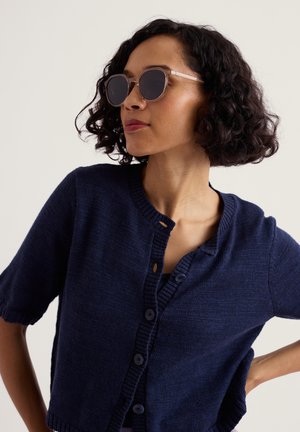 Femeie cu păr creț poartă ochelari de soare rotunzi cu ramă maro și un cardigan bleumarin cu mâneci scurte și nasturi, privind în lateral cu mâna pe șold.