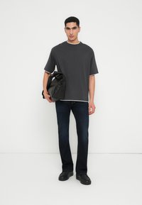 Jonge man met een donker oversized t-shirt en donkere jeans, met een zwarte leren tas in zijn hand, staand tegen een plain witte achtergrond.