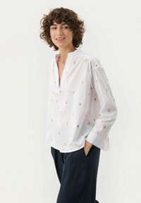 Valkoinen, pitkähiha-aineshirt, jossa on V-kaula-aukko ja kultaiset timanttikuvioinnit. Pehmeä puuvillakankaasta valmistettu, rento malli ja laskeutuvat hartiat. Yhdistetty tummiin housuihin.