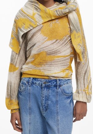Person wearing a yellow, beige, and gray patterned knit sweater with matching knit over-the-shoulder detail and blue high-waisted denim jeans.