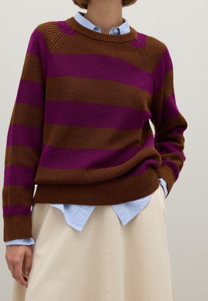 Personne portant un pull en maille rayé marron et violet par-dessus une chemise bleu clair à col avec une jupe couleur crème.