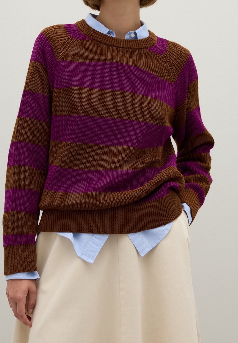 Personne portant un pull en maille rayé marron et violet par-dessus une chemise bleu clair à col avec une jupe couleur crème.