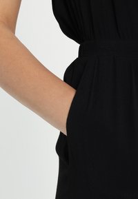 Černý oděv vyrobený z jemné, splývavé látky s nařaseným pasem a boční kapsou, zachycující detailní záběr paže odpočívající na bocích.