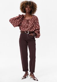 Brązowa blouse w kropki z bufiastymi rękawami i wiązaniem przy dekolcie, zestawiona z ciemnobrązowymi spodniami w wysokim stanie i wzorzystymi szpilkami.