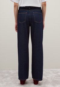 Jeans de mezclilla azul oscuro de pierna ancha con una cintura alta, que presentan costuras doradas en contraste y dos bolsillos traseros. Textura suave y de largo completo.