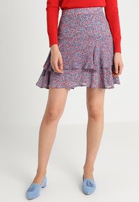 Femme portant une jupe à volants florale bleue, un haut rouge à manches longues et des mocassins à pampilles bleu clair, se tenant devant un fond blanc uni.