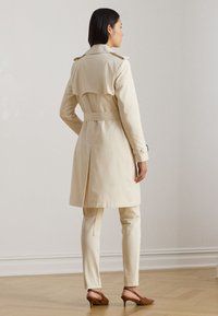 Beige trench coat with a belted waist, double-breasted design, and shoulder epaulettes; paired with beige pants and brown heeled shoes.