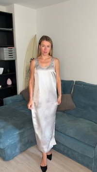 Femme debout à l'intérieur portant une longue robe en satin argenté et des talons noirs à côté d'un canapé turquoise et d'une planche de surf appuyée contre le mur.