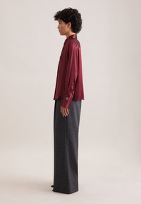Burgunderfarbige Satinbluse mit hohem Kragen, langen Ärmeln und dekorativen Knopfdetails, kombiniert mit hochtaillierten, maßgeschneiderten grauen Hosen.