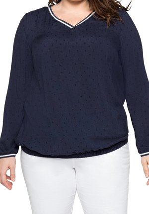 Femme portant un chemisier texturé bleu marine à manches longues avec un col en V rayé blanc et un pantalon blanc, debout avec les bras détendus le long du corps.