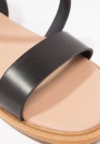 Sandale en cuir synthétique noir avec un design à une seule bride. Dispose d'une semelle intérieure lisse beige et d'une semelle flexible tan.