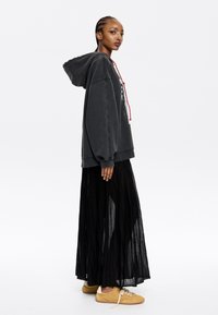 Felpa nera oversize con grafica bianca e cordini rossi, abbinata a una gonna maxi plissettata nera e scarpe in suede color beige.