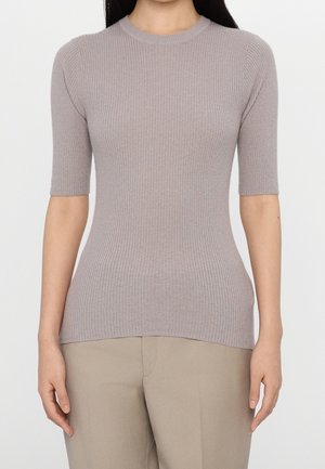 Femme portant un pull court à manches courtes, ajusté, côtelé, gris clair et un pantalon beige, debout sur un fond blanc uni.