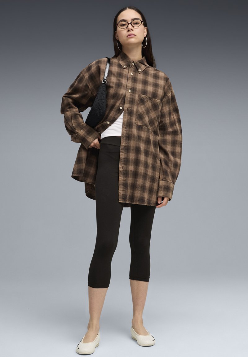 Femme portant une chemise à carreaux marron oversize, des leggings noirs courts, des chaussures blanches, un sac à bandoulière noir et des lunettes rondes, sur fond gris.