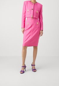 Completo sartoriale rosa con giacca cropped strutturata, dettagli in bottone dorato, due tasche a toppa e una gonna a tubino fino al ginocchio. Abbinato a tacchi viola.