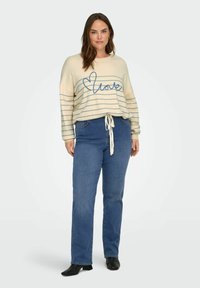 Femme portant un pull crème à rayures bleues avec le texte "love", un jean bleu avec un cordon blanc, et des bottines noires, debout devant un fond uni.