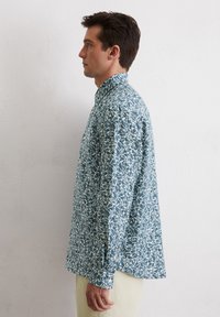 Chemise à manches longues bleu clair avec un motif de feuilles blanches, poignets boutonnés et col, fabriquée en tissu doux et texturé.