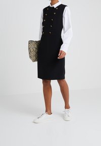 Musta ilmanhiha-haalari, jossa on kultaisia nappeja, yhdistettynä valkoiseen paitaan ja valkoisiin tennareihin. Pitelee vaaleaa käärmeennahkakuosisia käsilaukkua.