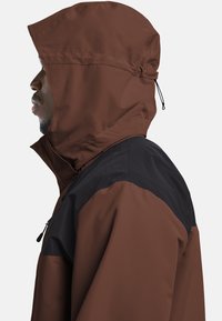 Braune wasserdichte Jacke mit schwarzem Schulterpanel, hohem Kragen und angeschnallter Kapuze. Verfügt über einen elastischen Kordelzug und Reißverschlusstaschen.