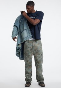 Jeansjacke in Blautönen, leicht ausgebleicht; marineblauses T-Shirt; olivgrüne Camouflage-Cargo-Hose mit Taschen; dunkle Sneakers. Schlichte, funktionale Stilrichtung.