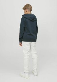 Felpa con cappuccio blu scuro, dotata di un ampio cappuccio, con polsini e fondo a costine, abbinata a pantaloni della tuta grigi chiari. Modello di spalle, che indossa sneaker bianche.