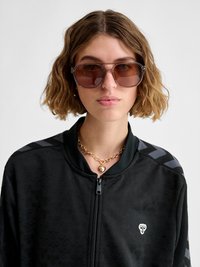 Giovane donna con capelli castani ondulati che indossa grandi occhiali da sole marroni, una collana con ciondolo a forma di cuore in oro e una giacca nera con zip con un piccolo logo bianco.