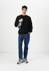 Felpa nera, jeans blu e sneakers bianco e neri. Un marsupio con motivo si trova sul lato sinistro, dotato di chiusura con cerniera.