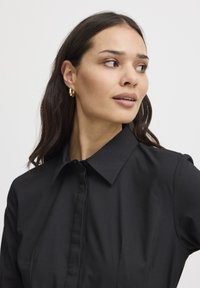 Camicia nera con colletto, tessuto liscio e chiusura a bottoni. La modella indossa orecchini a cerchio dorati, mostrando un'espressione neutra.