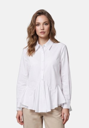 Donna con capelli ondulati che indossa una camicia bianca a maniche lunghe con bottoni e dettagli plissettati sull'orlo e pantaloni beige su uno sfondo semplice.