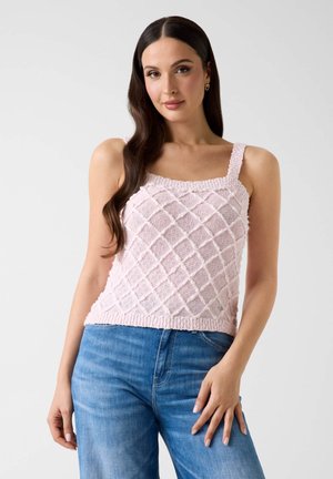 Femme aux longs cheveux bruns portant un débardeur en maille texturée rose clair et un jean bleu sur un fond uni.