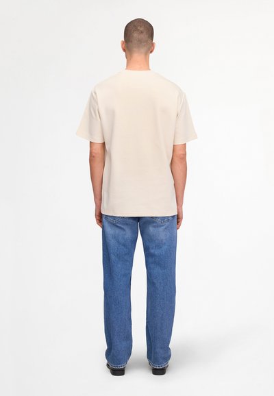 Ein Mann steht mit dem Rücken zum Betrachter und trägt ein schlichtes, beiges T-Shirt mit kurzen Ärmeln, blaue Jeans und schwarze Schuhe vor weißem Hintergrund.