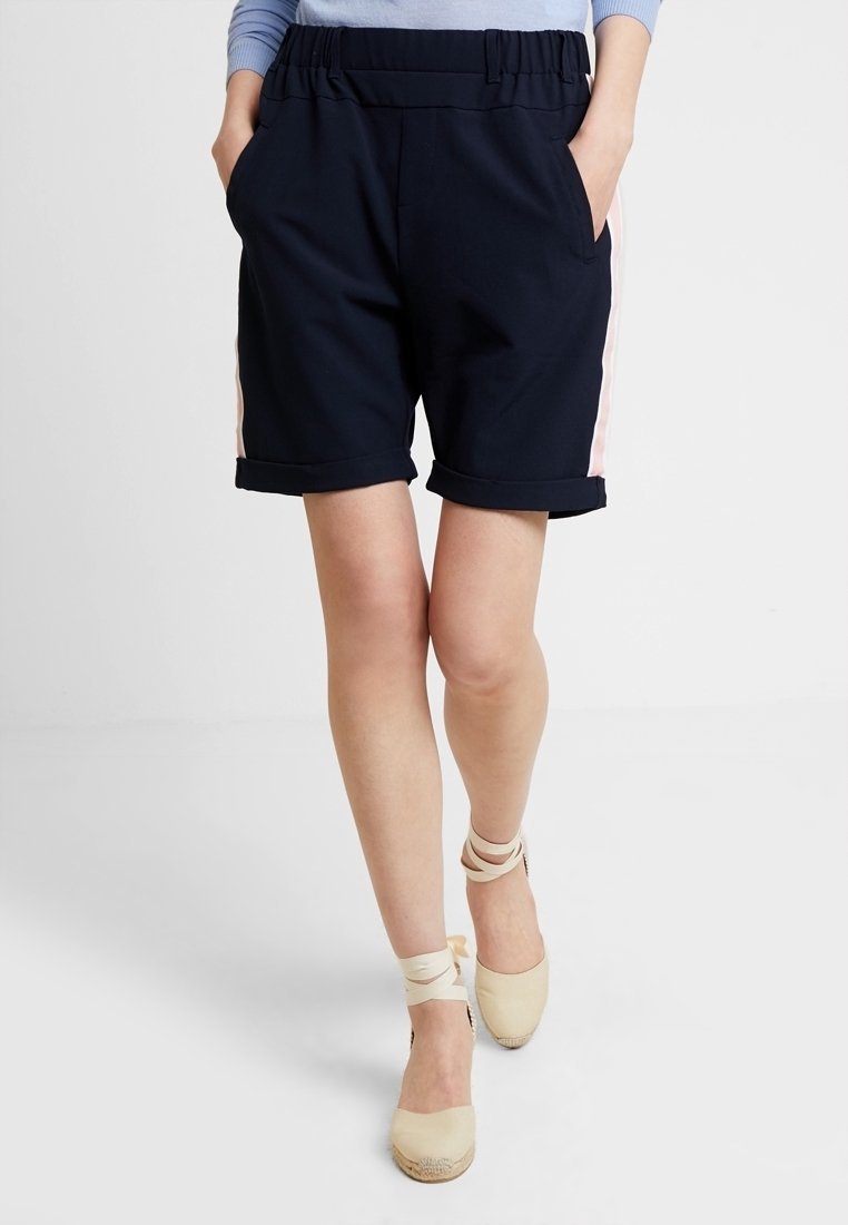 Femme portant un short bleu marine avec des bandes latérales roses et des espadrilles beige, les mains dans les poches, debout devant un fond uni.