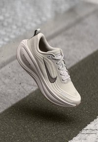Light beige Nike running shoe with ZoomX sole midair above textured pavement with white road markings.