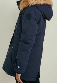 Navy blue insulated parka with a faux fur hood, zip closure, side pockets, and a logo patch on the arm. Soft texture and mid-length design.