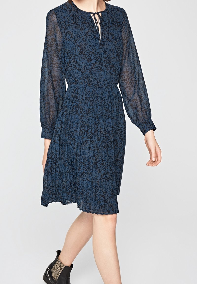 Robe bleue à motifs longueur genoux avec manches longues transparentes, col noué, jupe plissée, portée avec des bottines noires avec un détail imprimé serpent.