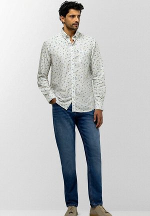 Mann mit kurzen lockigen Haaren, der ein weißes langärmliges gemustertes Hemd mit Knöpfen, blaue Jeans und beige Schuhe trägt, steht vor einem schlichten Hintergrund.