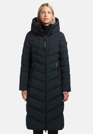 Woman wearing a long, black, quilted winter coat with a hood, front zipper, and zippered pockets, standing against a plain background.