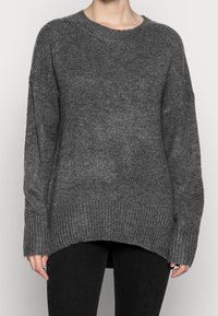 Person, der einen lockeren dunkelgrauen Strickpullover mit gerippten Bündchen und Saum trägt, kombiniert mit schwarzen Hosen, vor einem schlichten Hintergrund steht.