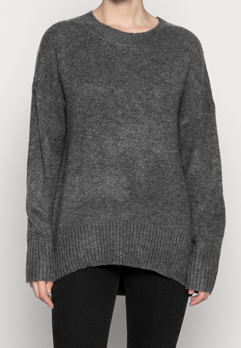 Person, der einen lockeren dunkelgrauen Strickpullover mit gerippten Bündchen und Saum trägt, kombiniert mit schwarzen Hosen, vor einem schlichten Hintergrund steht.