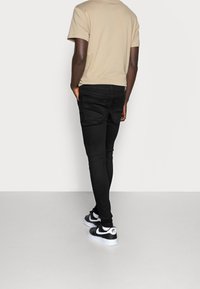 Beige bomulls-t-shirt, svarta tajta jeans, svartvita sneakers, sedd bakifrån. Mjuk textur, enkel design, inga synliga mönster.