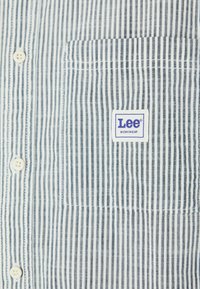 Striped button-up shirt in white and navy, made of lightweight fabric. Features a chest pocket with a Lee Workwear logo patch.