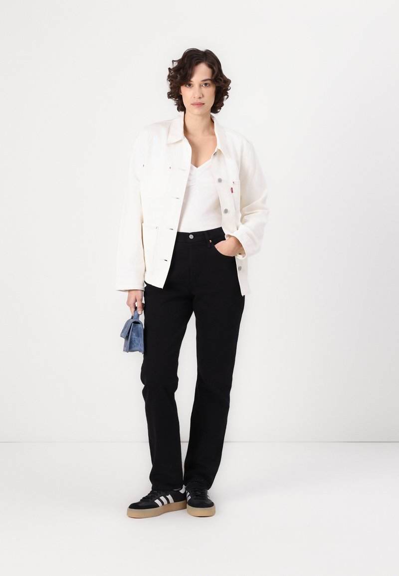Mujer de pie con una mano en el bolsillo de los pantalones negros, vistiendo una chaqueta y blusa blancas, zapatillas negras, y sosteniendo un pequeño bolso azul.