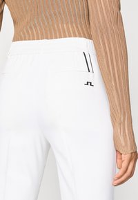 Baltas bikses ar strukturētu jostu, kurām ir divas aizmugurējās kabatas un melna logo. Audums šķiet gluds un viegls.