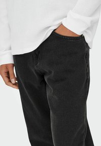 Jeans de mezclilla negra con un corte recto, costuras visibles y una ligera textura. Combinados con una camiseta blanca de manga larga con textura.