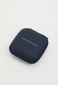 Tamsiai mėlyna užtrauktuko dėžutė su lygiu, švelniu paviršiumi. Puošia balta emblema su užrašu "TOMMY HILFIGER." Kompaktiška kvadratinė forma, be papildomų puošmenų.