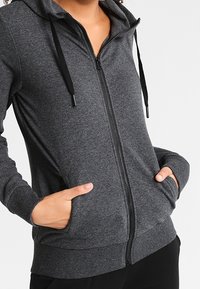 Sudadera con capucha gris con cremallera, con cordón en la capucha, bolsillos delanteros y puños acanalados. Textura suave y ajuste casual. Cremallera en la parte delantera central.